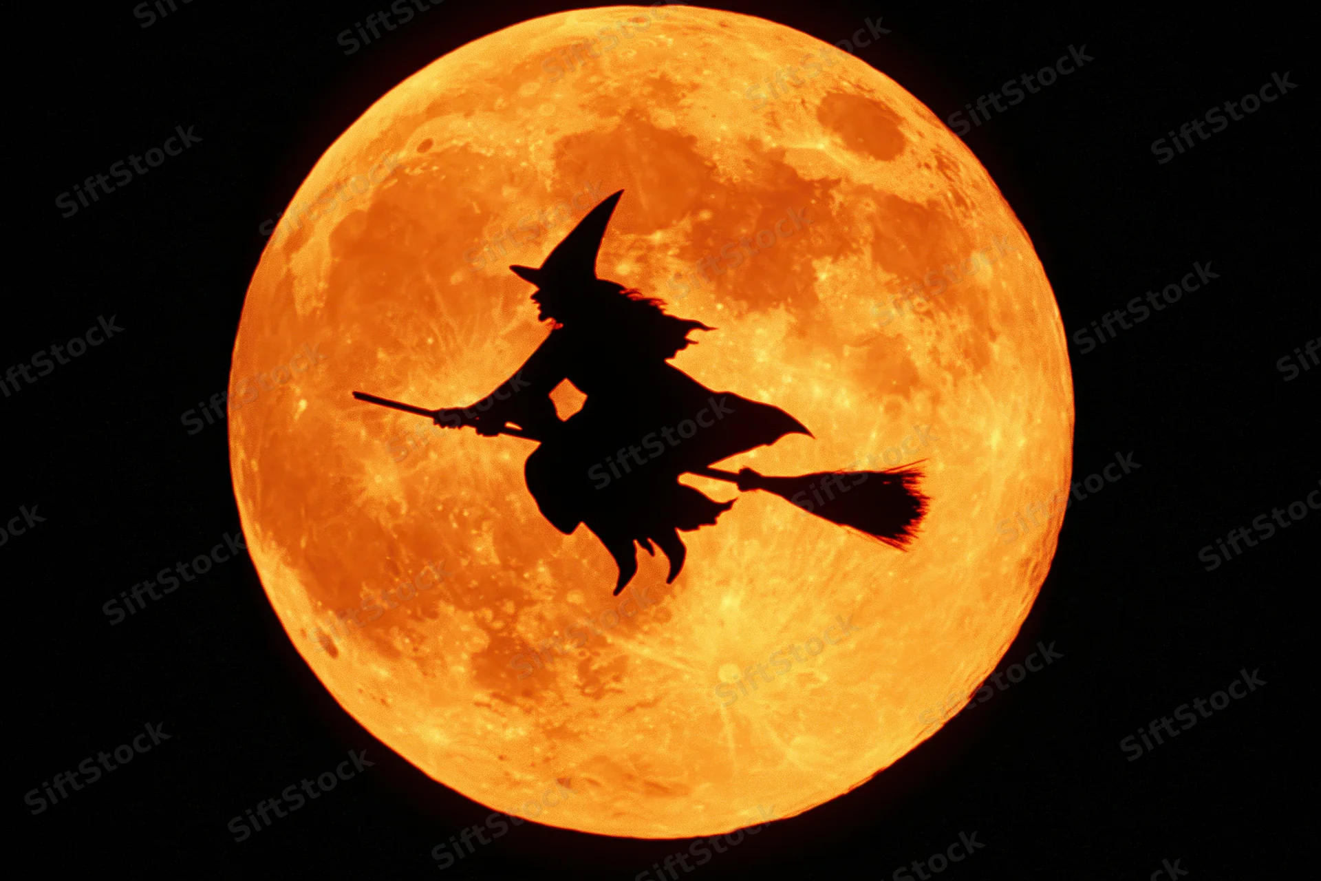 Silhouette of a witch riding a broomstick in flight centered against a glowing orange full moon at night
