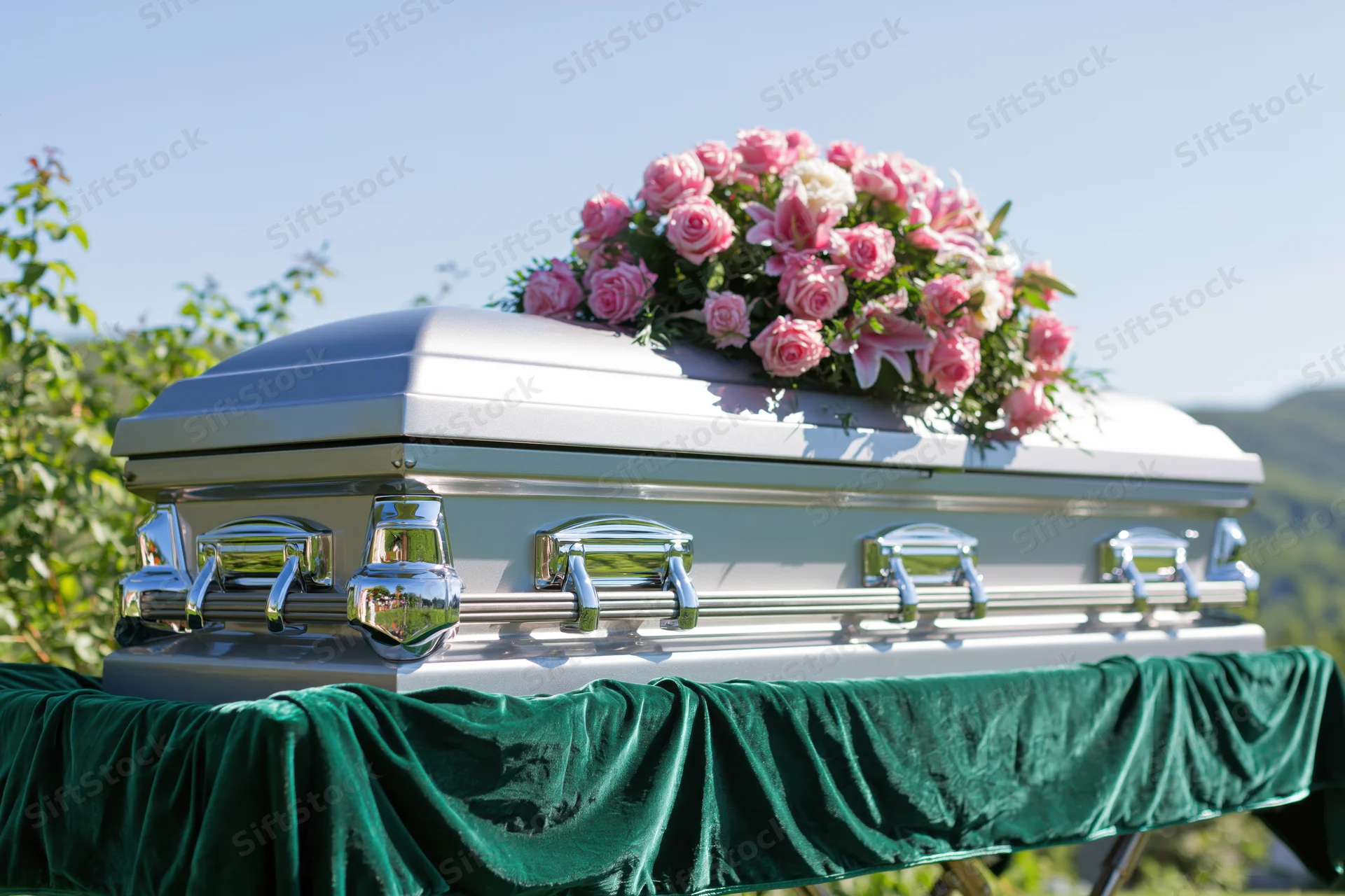 Silver metallic casket with pink roses and lilies on a green velvet-draped bier outdoors
