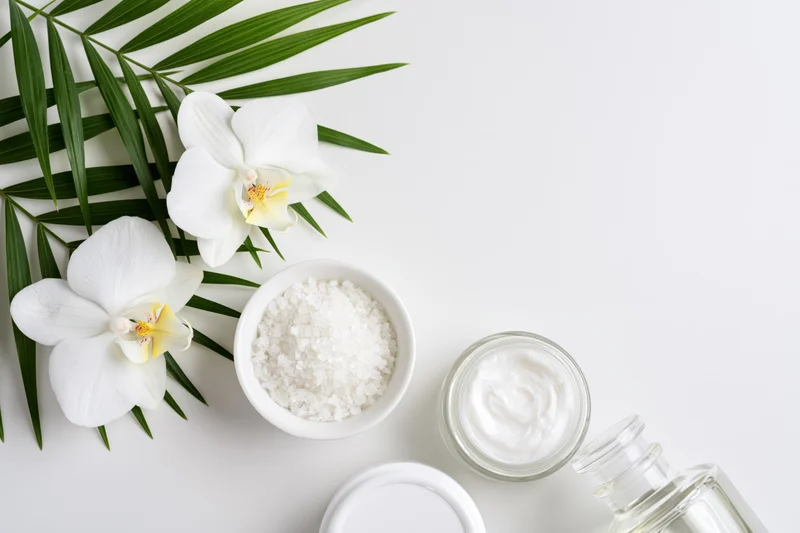 Flatlay of skincare products with white orchids and green leaves on white surface