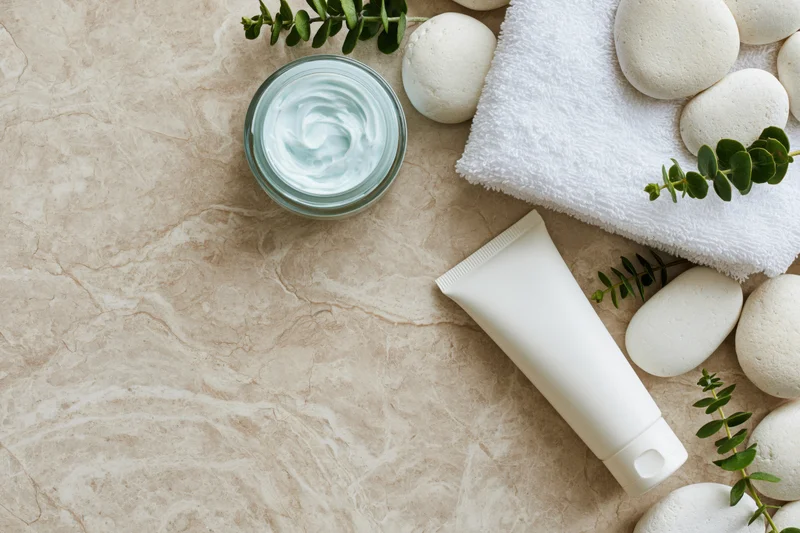 Open glass jar of pale blue cream and white tube with towel, stones and eucalyptus on marble surface