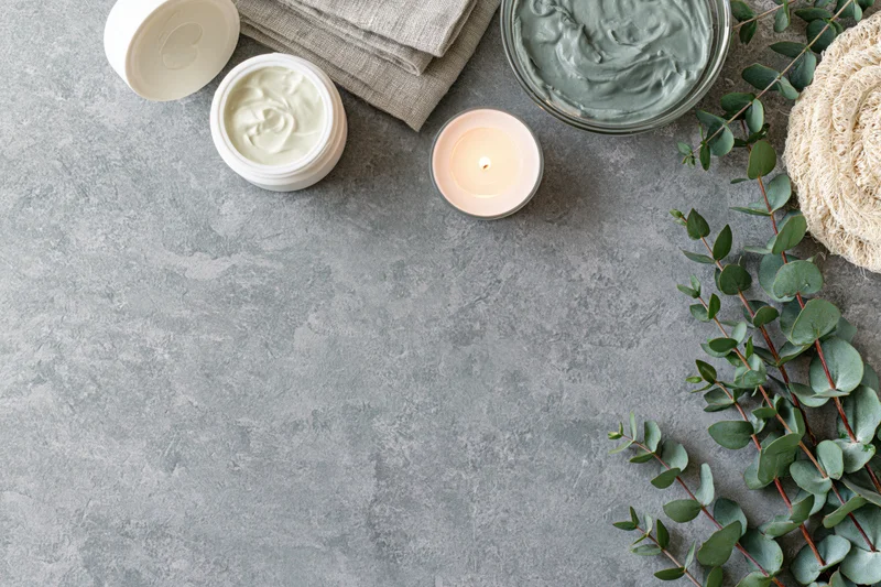 Open cream jar and lit candle with eucalyptus on gray surface in flatlay arrangement