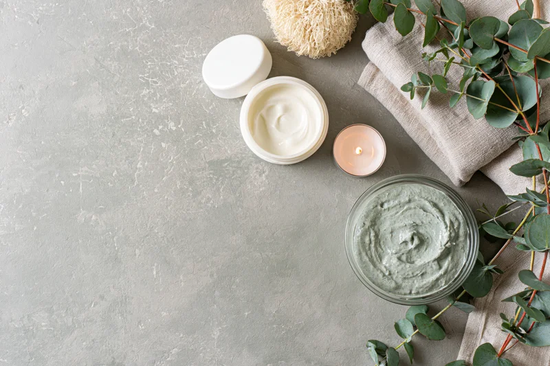 Open white cream jar and green facial mask with eucalyptus and candle on gray surface