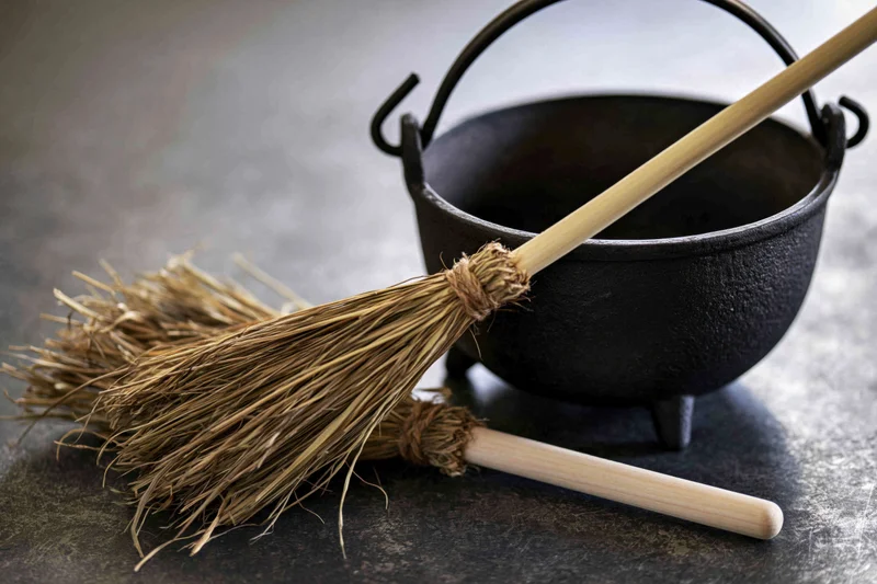 Small black cast iron cauldron with a broom resting beside it on a dark textured surface