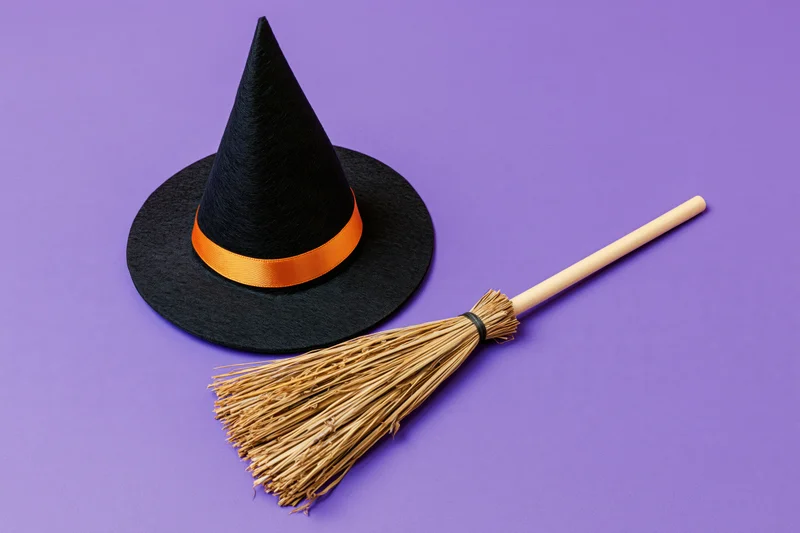 Black witch hat with orange ribbon and straw broom on purple background