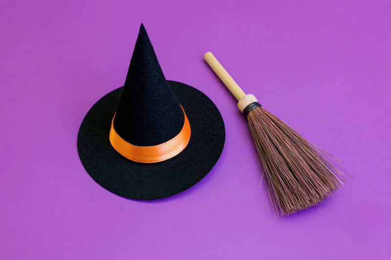 Black witch hat with orange ribbon and small broom on purple background