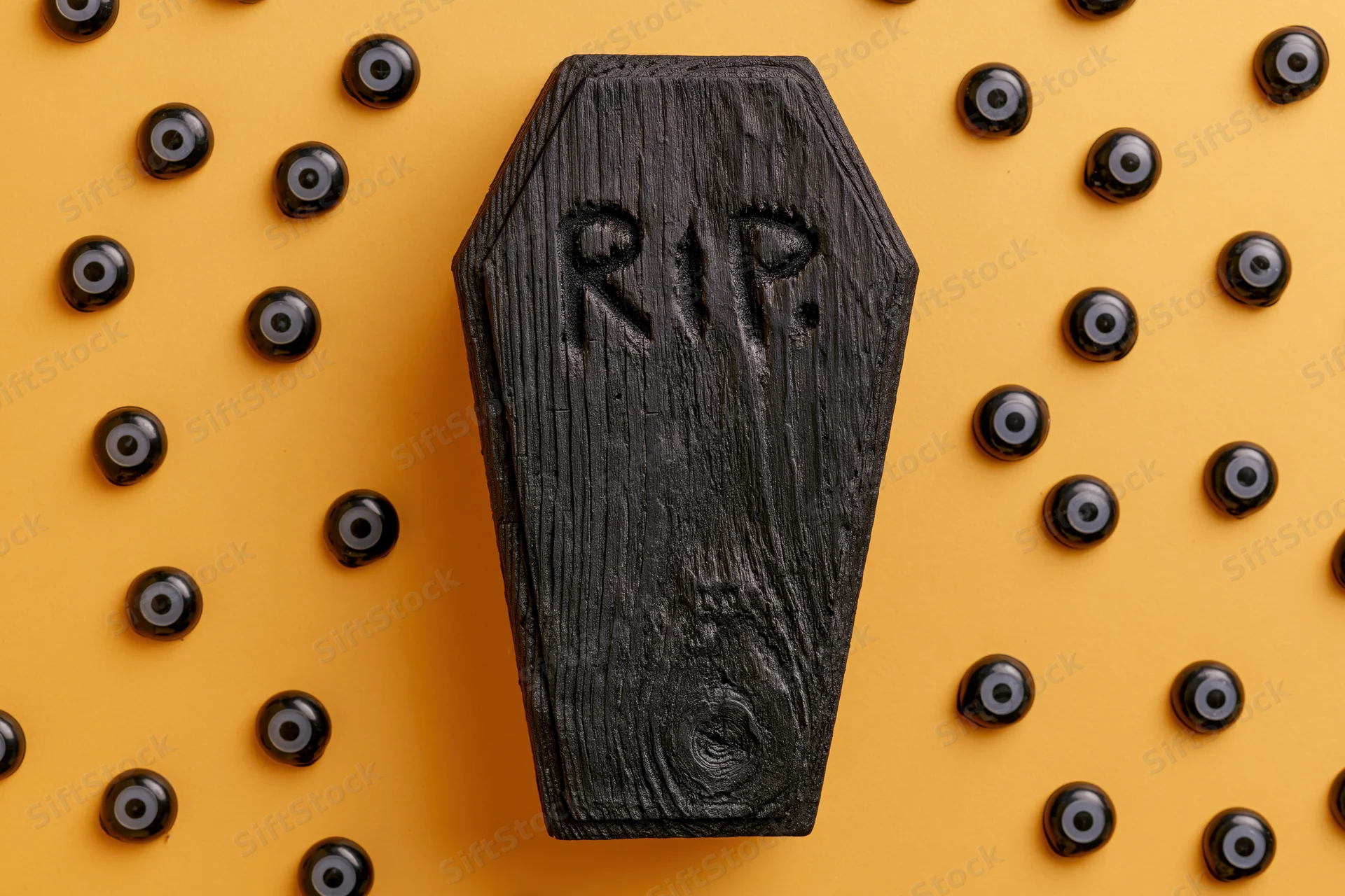 Small black wooden coffin with rip carved on it surrounded by black eyes on orange background