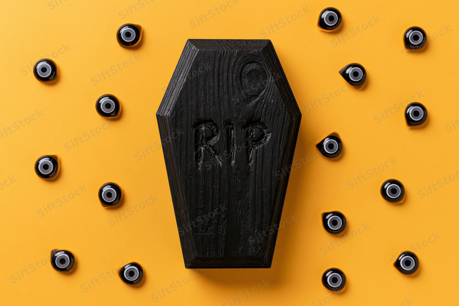 Small black wooden coffin with rip inscription surrounded by plastic eyeballs on orange background