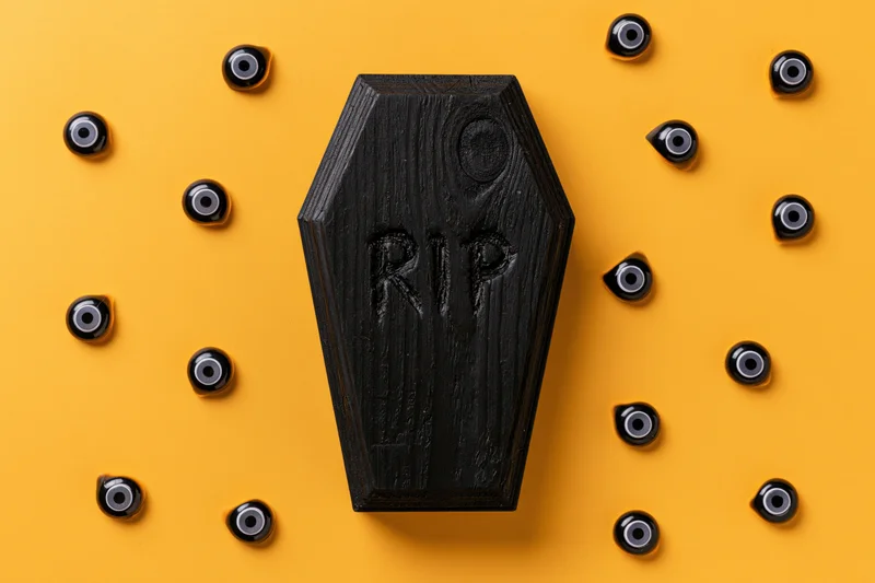 Small black wooden coffin with rip inscription surrounded by plastic eyeballs on orange background