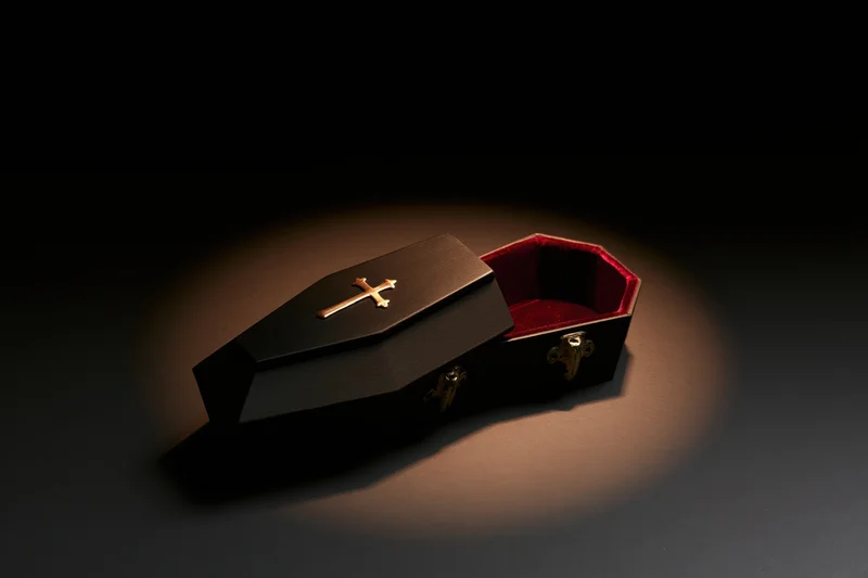 Small dark wooden coffin model with open lid and gold cross under spotlight on black background