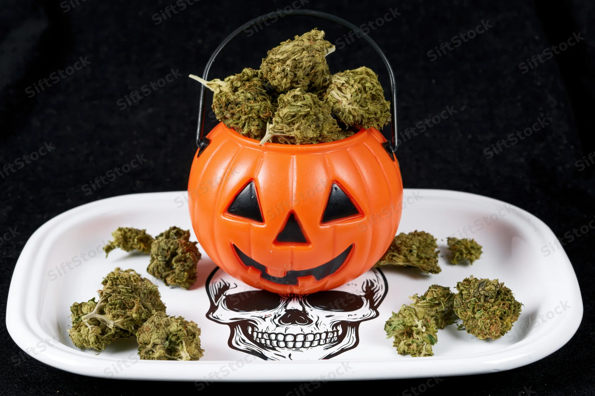 Orange jack-o-lantern bucket filled with cannabis buds on a white tray with skull design, set against dark background