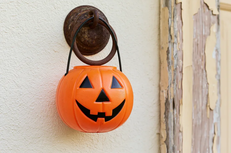 Orange plastic jack-o-lantern candy bucket hanging on a rusty door knocker beside a peeling wooden frame