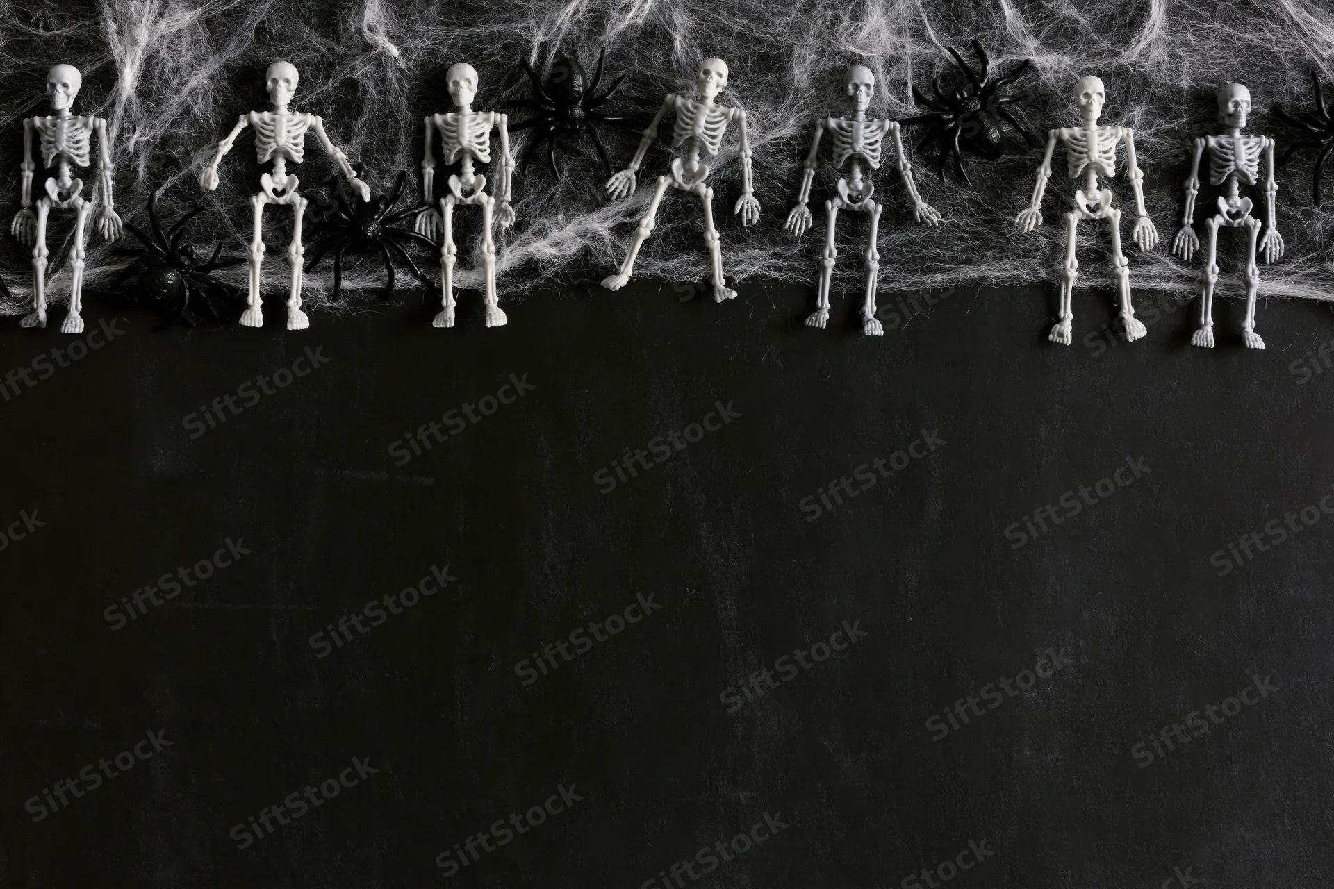 Small plastic skeleton figurines and black spiders arranged in a row on a black surface with cobwebs above, creating a Halloween decoration display