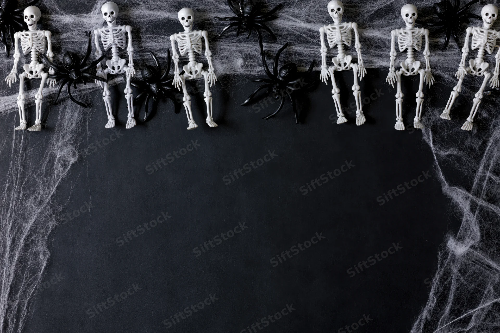 Small plastic skeletons and black spiders arranged in a row on a dark surface with cobwebs, creating a Halloween border with copyspace below