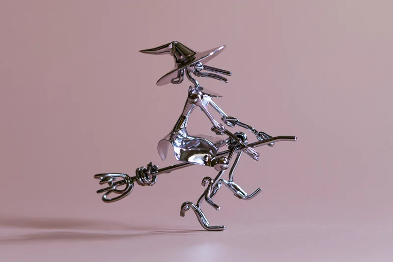 Chrome witch figurine riding a broomstick against a pink background