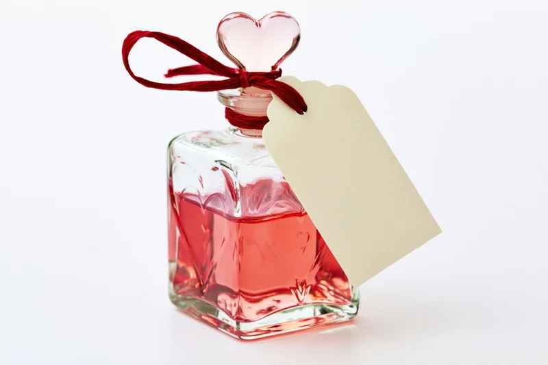 Square glass perfume bottle with heart-shaped stopper and red ribbon, holding pink-red liquid, attached to blank tag on white background