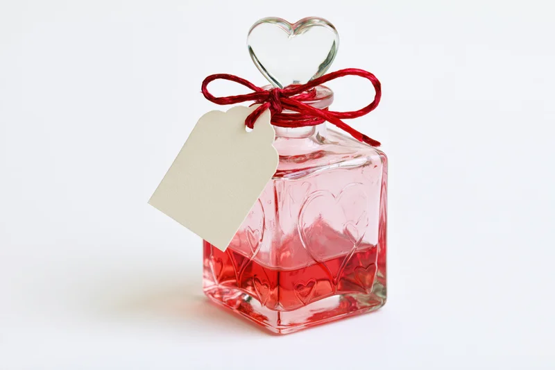 Square glass perfume bottle with heart-shaped stopper and red ribbon tied around neck, filled with pink-red liquid, white background