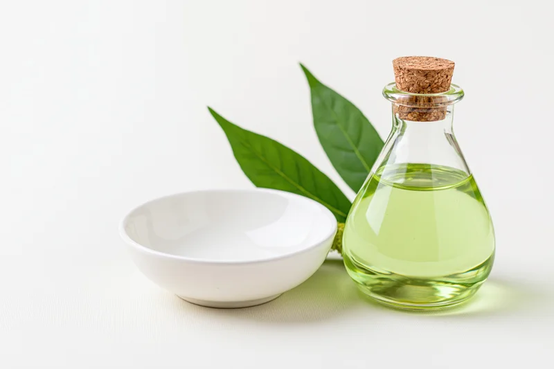 Glass bottle of green essential oil with cork stopper and fresh green leaves beside a white ceramic bowl on white surface