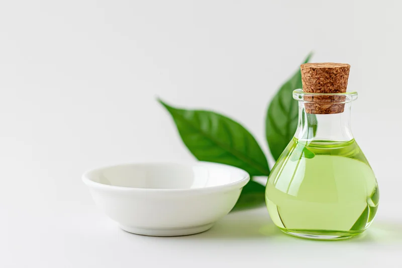 Glass bottle with green liquid and cork stopper beside a white ceramic bowl with green leaves on white background