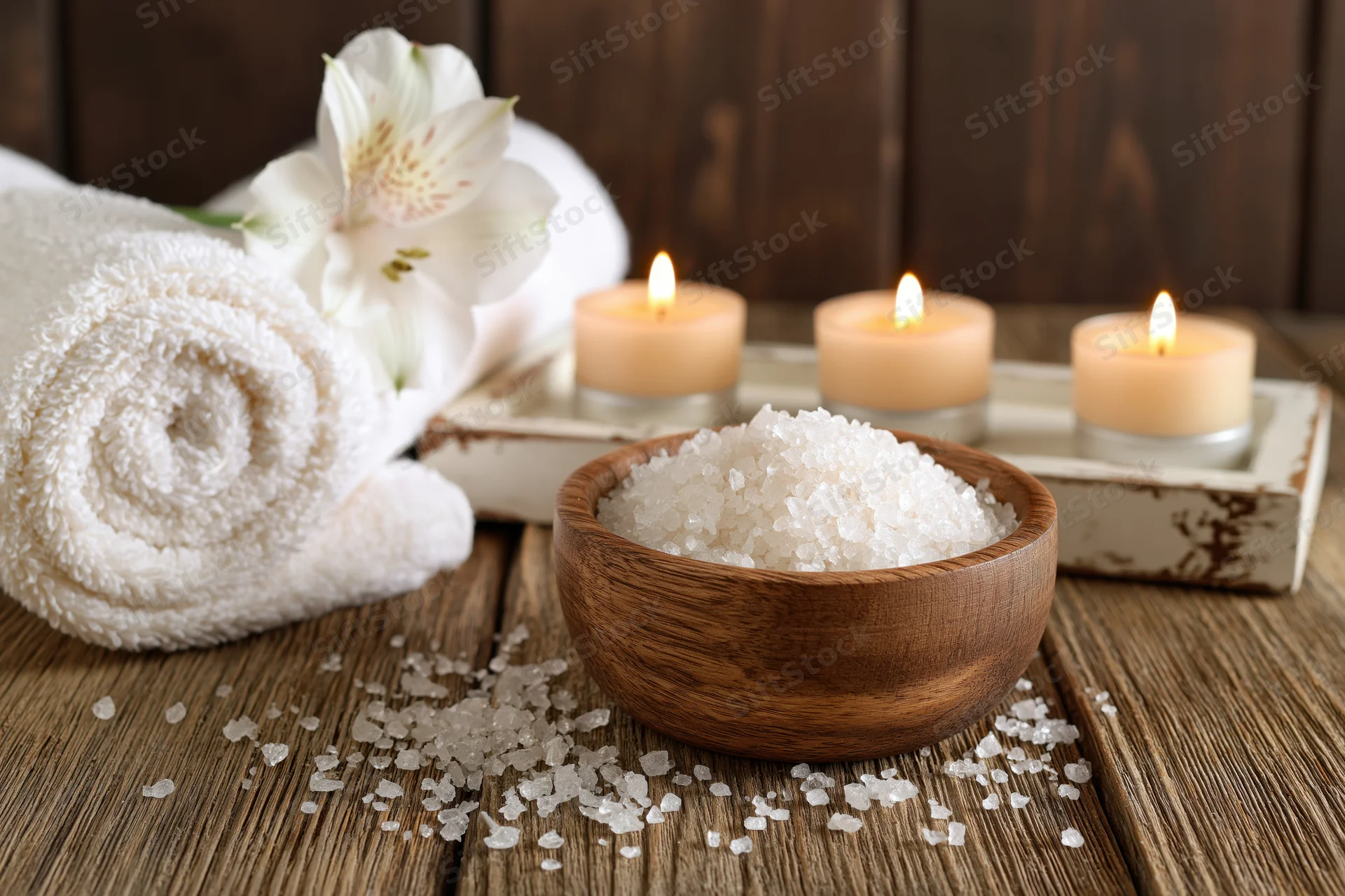Wooden bowl filled with coarse white salt on a rustic wooden table beside candles and a rolled towel