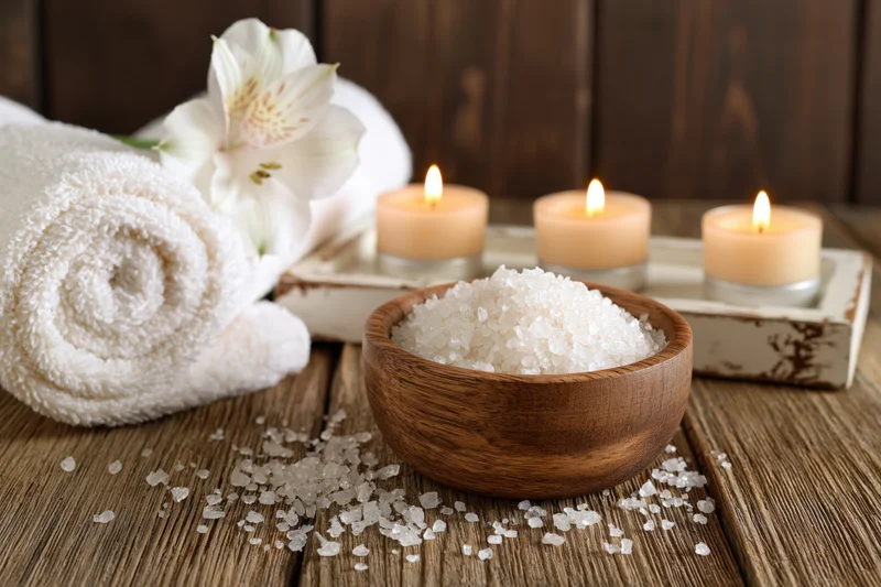 Wooden bowl filled with coarse white salt on a rustic wooden table beside candles and a rolled towel