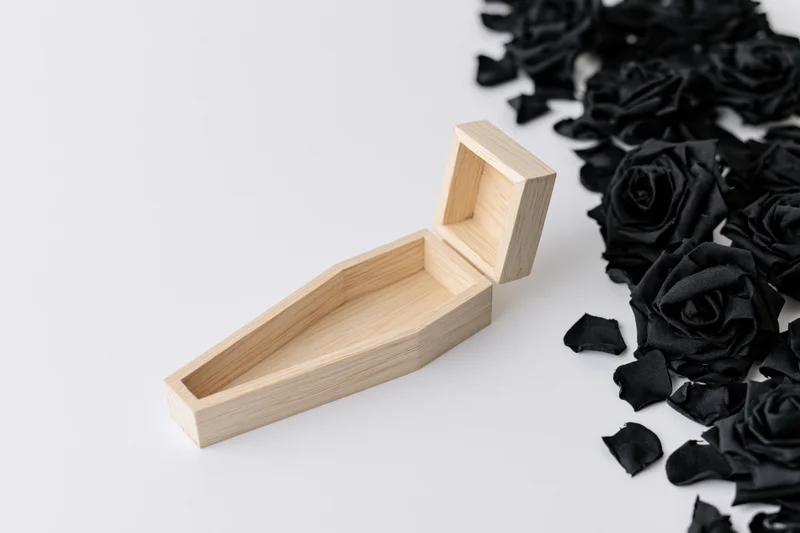 Small wooden miniature coffin with open lid lying flat beside black roses and petals on white surface