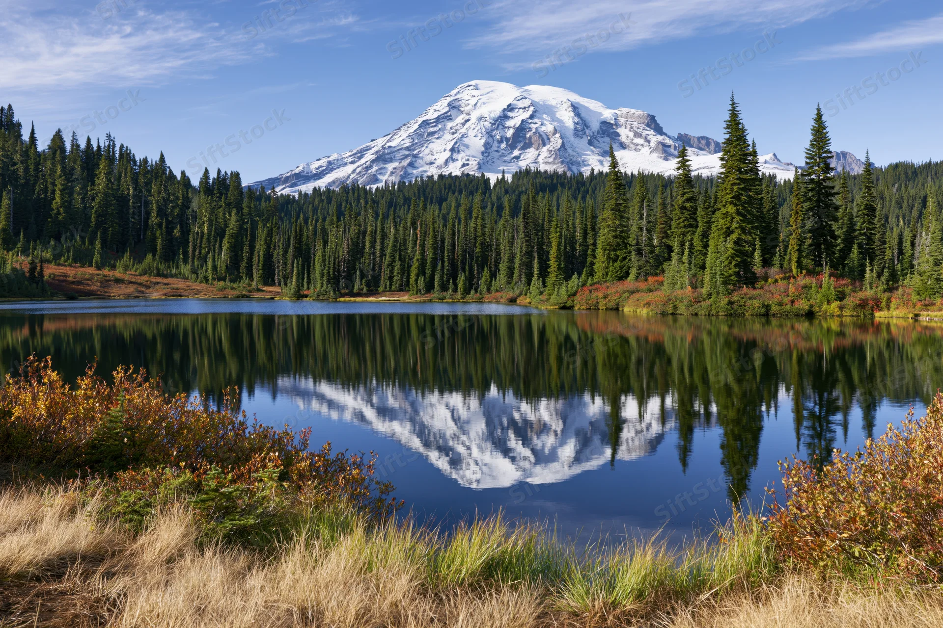 Snow-capped mountain reflected in calm lake surrounded by evergreen forest and autumn foliage