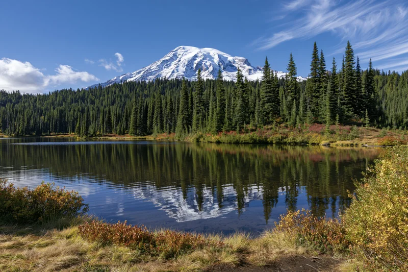Snow-capped mountain reflected in calm lake surrounded by evergreen forest and autumn foliage