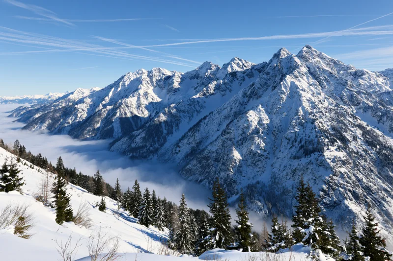 Snow-covered alpine mountains rising above a low cloud layer under a clear blue sky