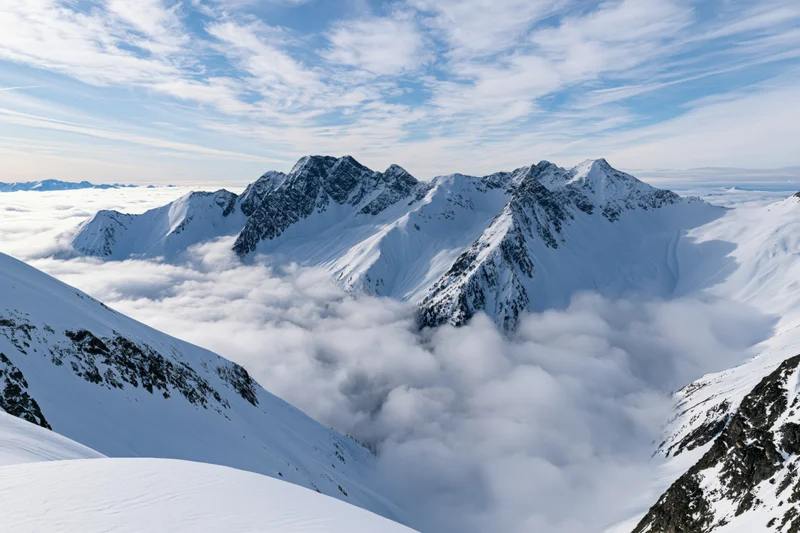 Snow-covered alpine mountain range rising above a low cloud layer under a blue sky