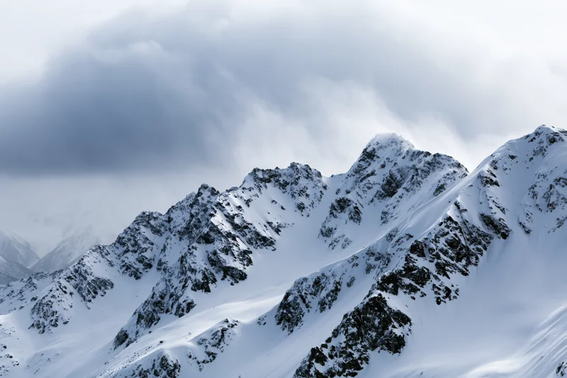 Snow-covered alpine mountain range with rocky ridgelines under cloudy sky