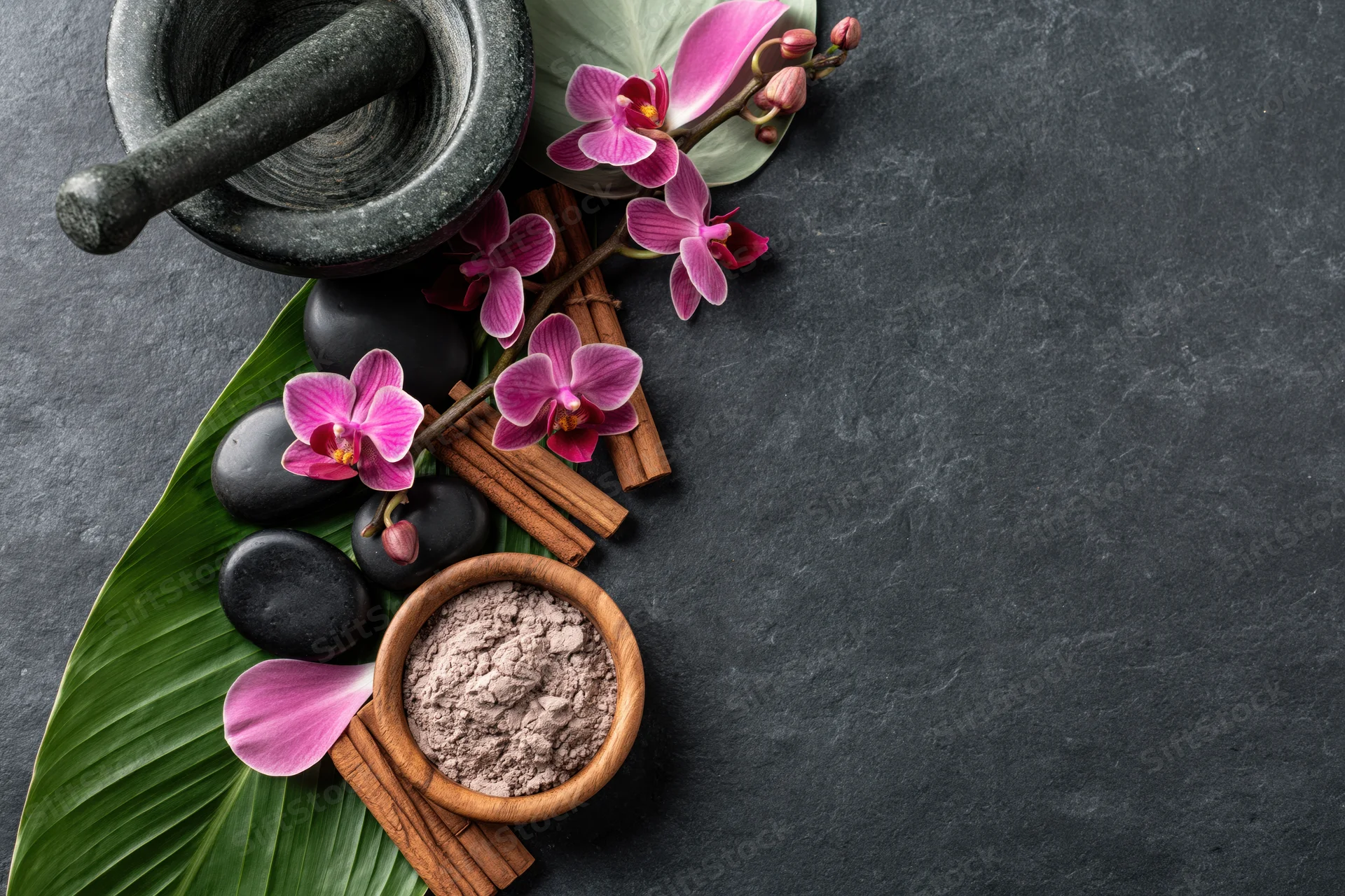 Spa arrangement with mortar, pestle, orchids, cinnamon sticks and stones on dark slate surface
