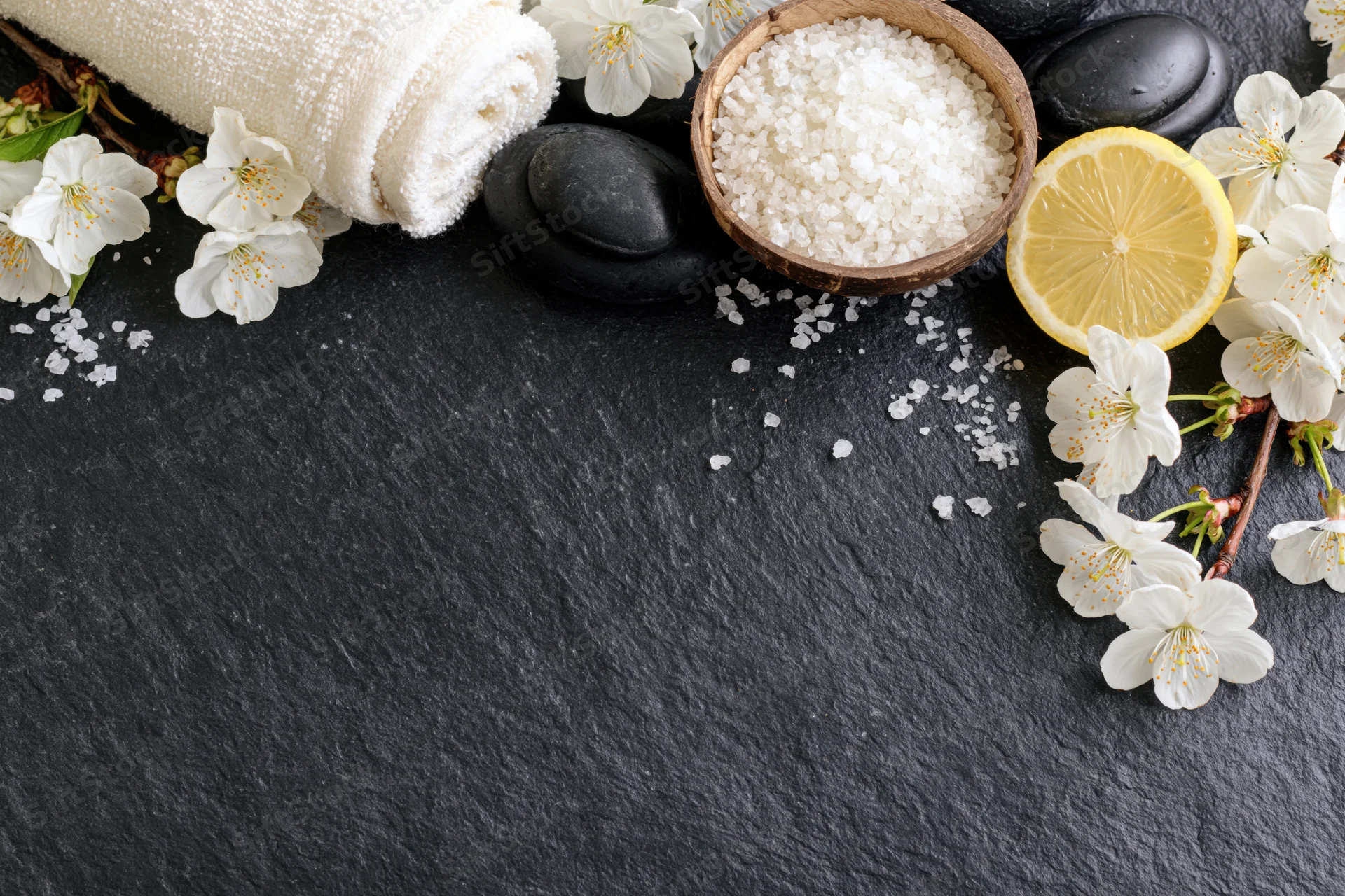 Spa flatlay arrangement with towel, flowers, lemon, salt and stones on dark slate surface