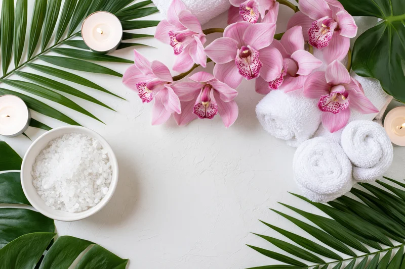 Pink cymbidium orchids and green leaves arranged with candles, towels, and salt on white surface for spa flatlay