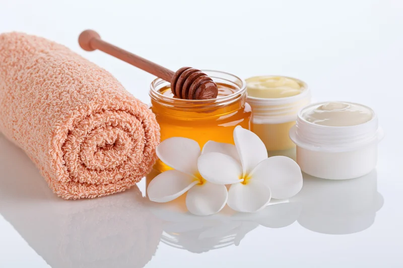 Rolled peach towel beside honey jar with wooden dipper, cream pots and frangipani flowers on reflective white surface