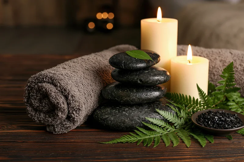 Stacked black stones with lit candles, fern, salt bowl and rolled towel on dark wooden surface