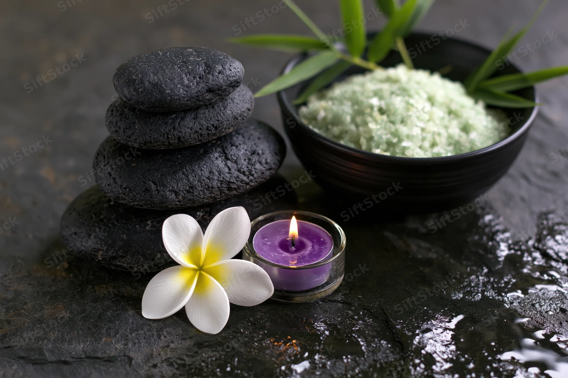 Stacked black stones, lit purple candle, white frangipani flower, and bowl of green salt on wet dark slate surface