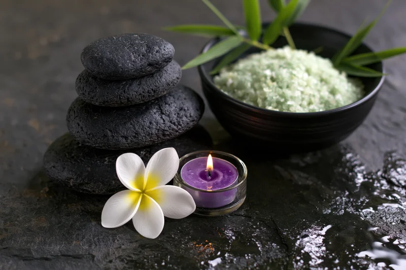 Stacked black stones, lit purple candle, white frangipani flower, and bowl of green salt on wet dark slate surface