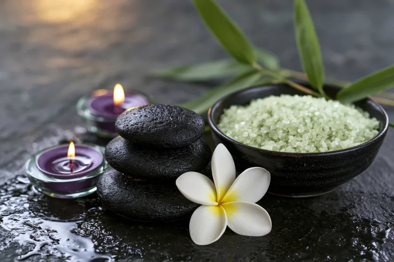 Spa still life with stacked stones, lit candles, salt bowl, frangipani flower and bamboo leaves on wet dark slate surface