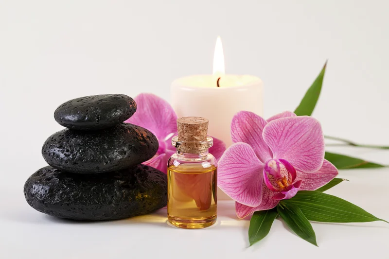 Stacked black stones, lit candle, essential oil bottle and pink orchid on white background