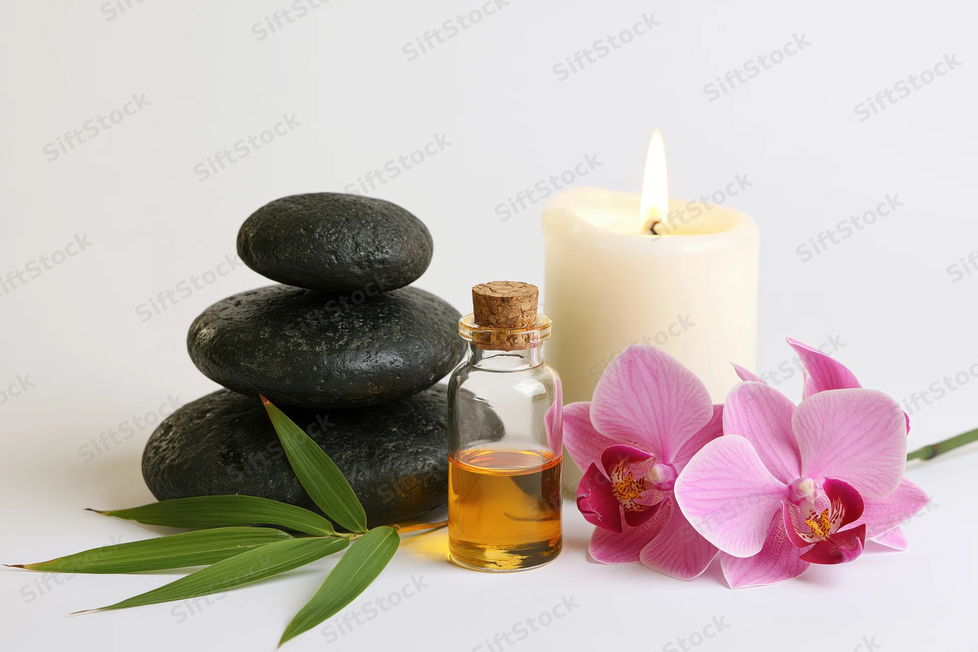 Spa still life with stacked black stones, essential oil bottle, lit candle, pink orchid and bamboo leaves on white background