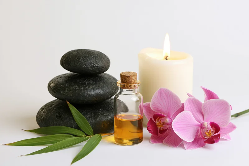 Spa still life with stacked black stones, essential oil bottle, lit candle, pink orchid and bamboo leaves on white background