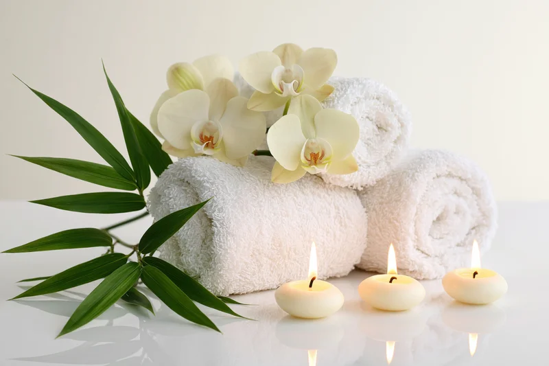 Rolled white towels with white orchids and bamboo leaves arranged beside three lit candles on a reflective white surface
