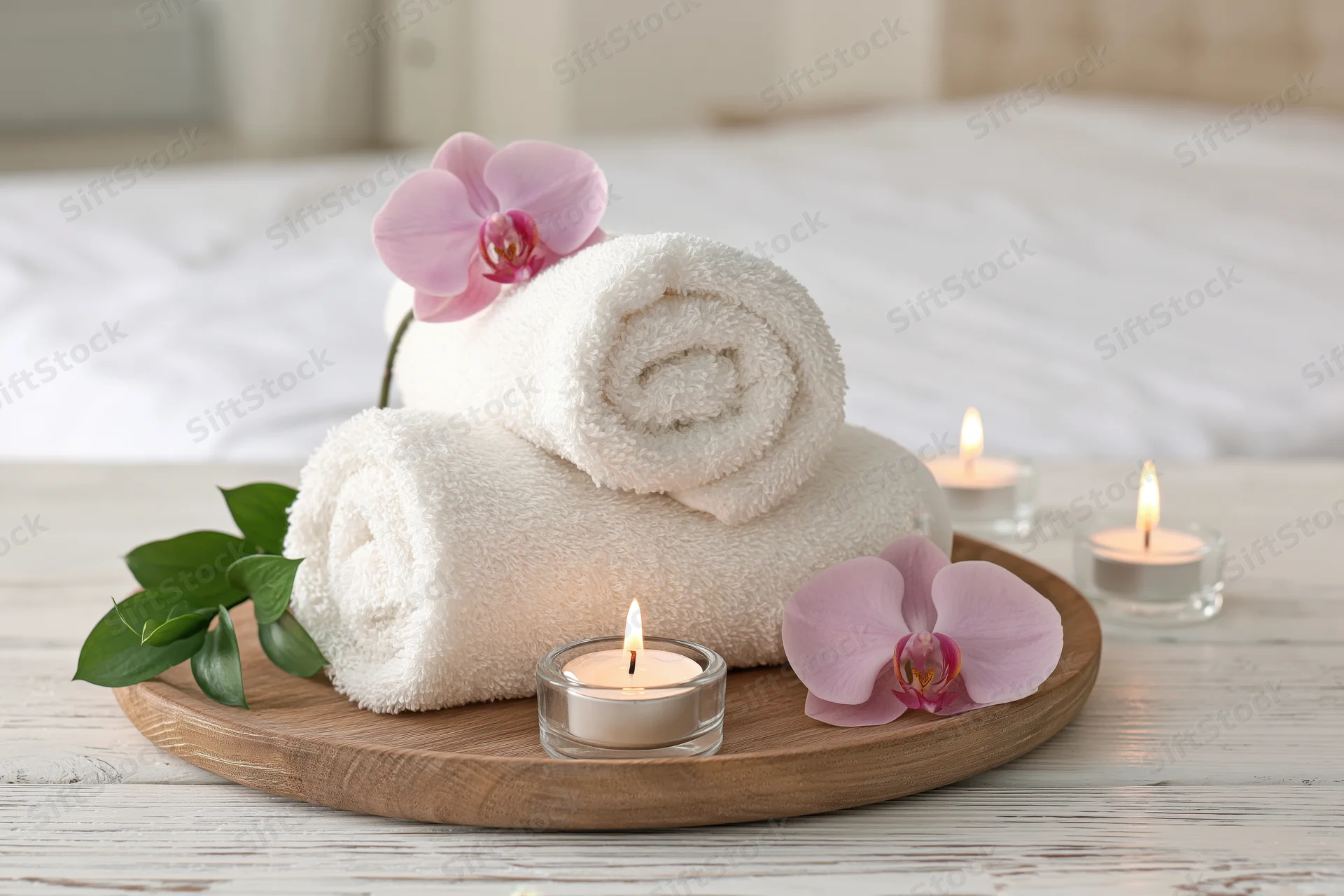 Rolled white towels with pink orchids and lit candles on a wooden tray beside a bed