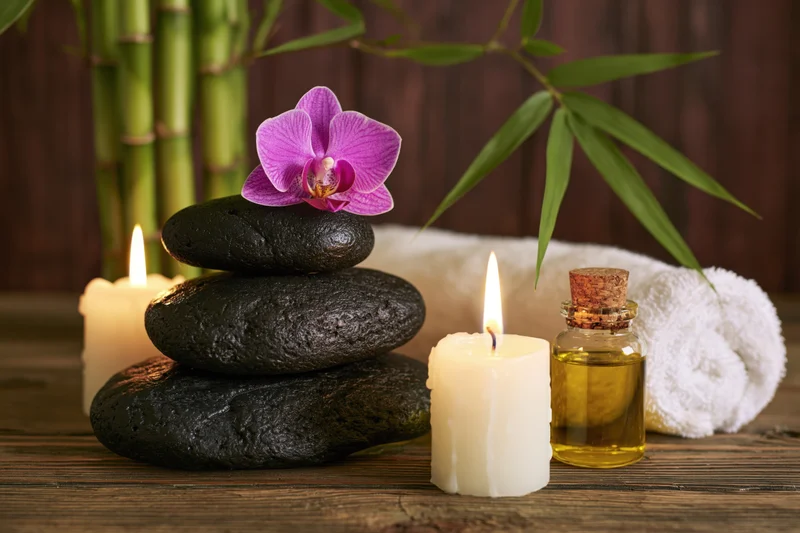Stacked black stones with a purple orchid, lit candles, essential oil bottle and white towel on a wooden surface