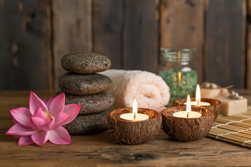 Spa still life with lit coconut candles, stacked stones, pink lotus, towel and bamboo mat on wooden surface