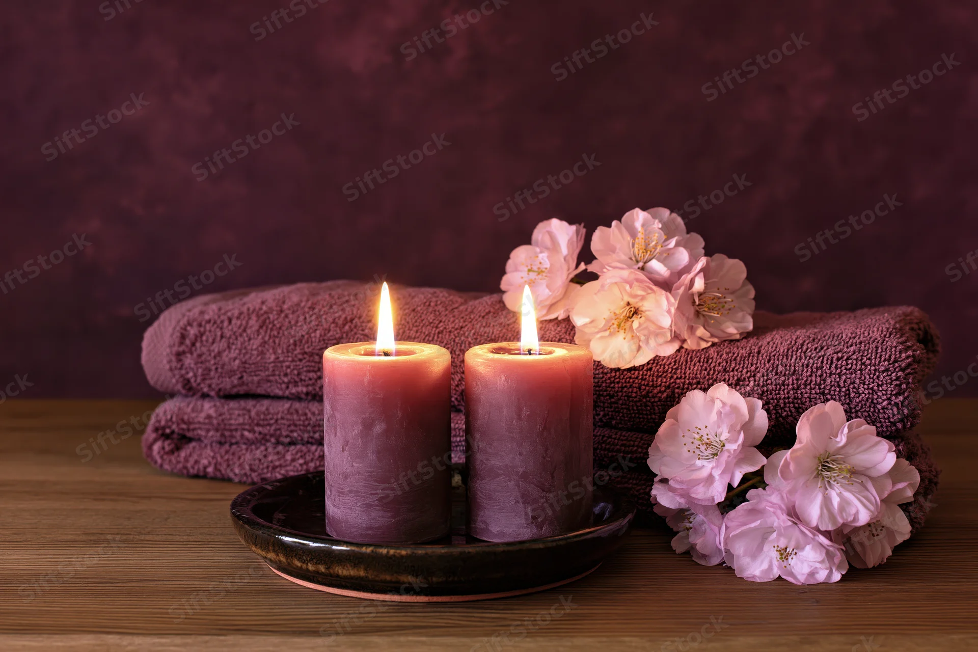 Two lit purple candles on a ceramic plate beside folded towels and pink cherry blossoms on a wooden surface