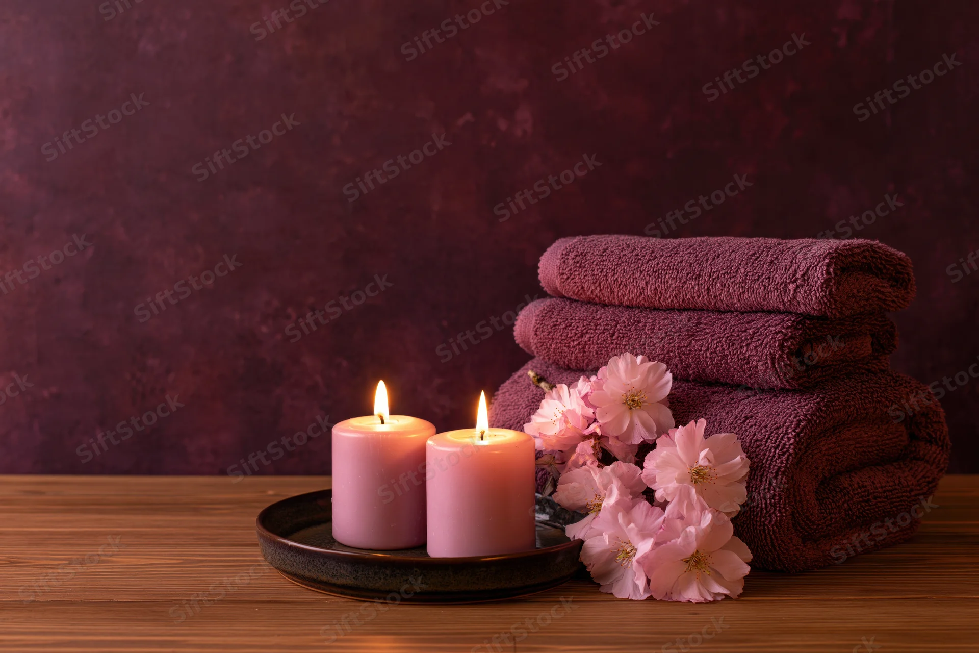 Two lit pink candles on a tray beside folded towels and cherry blossoms on a wooden surface