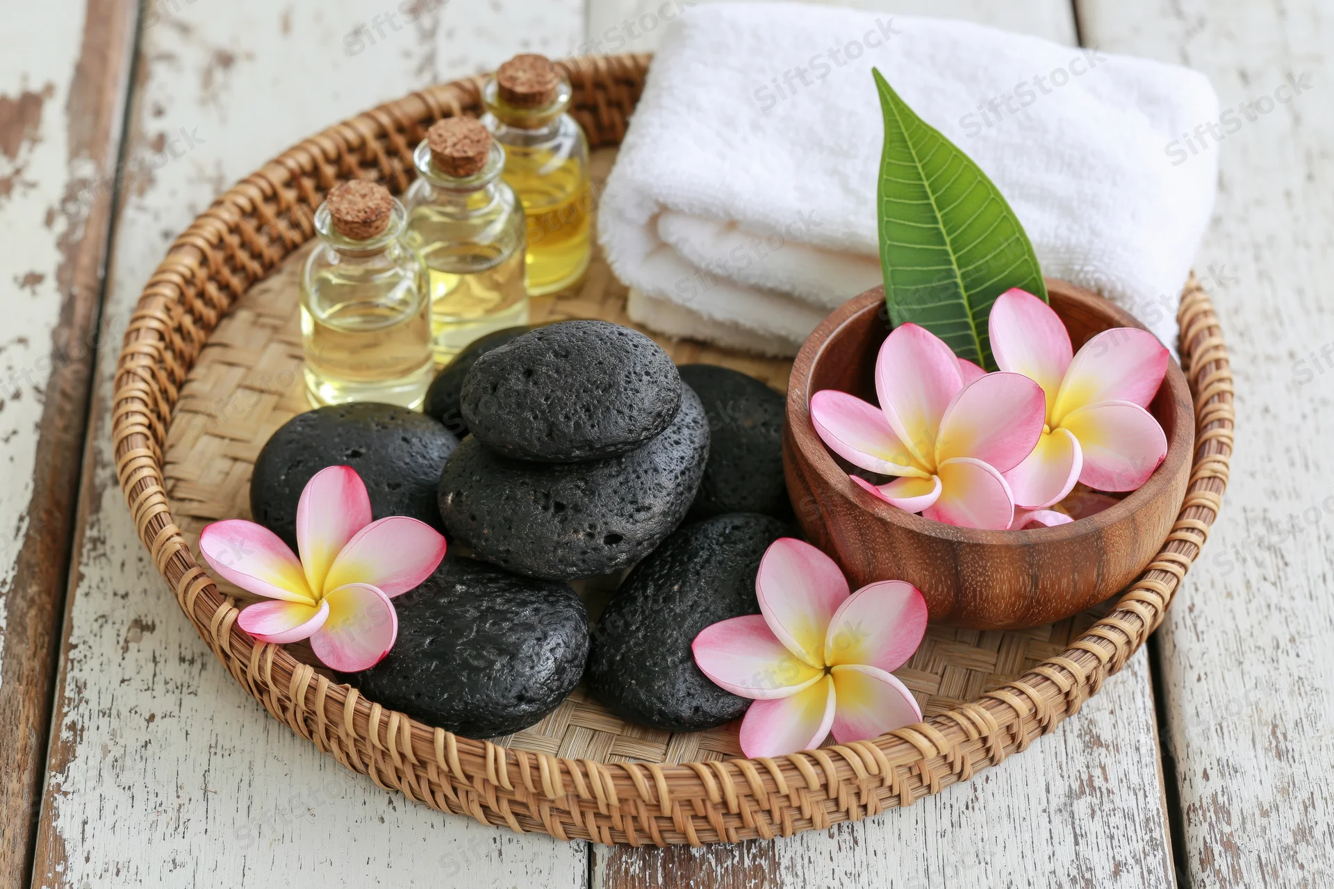 Spa still life with black stones, essential oil bottles, frangipani flowers and white towel arranged on a woven bamboo tray