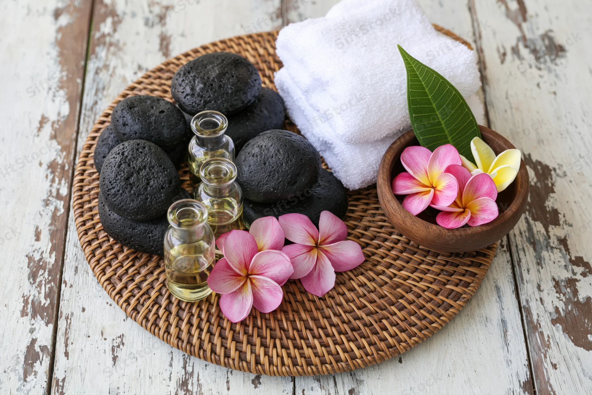 Spa still life with black stones, essential oil bottles, frangipani flowers, white towel and wooden bowl on woven bamboo tray