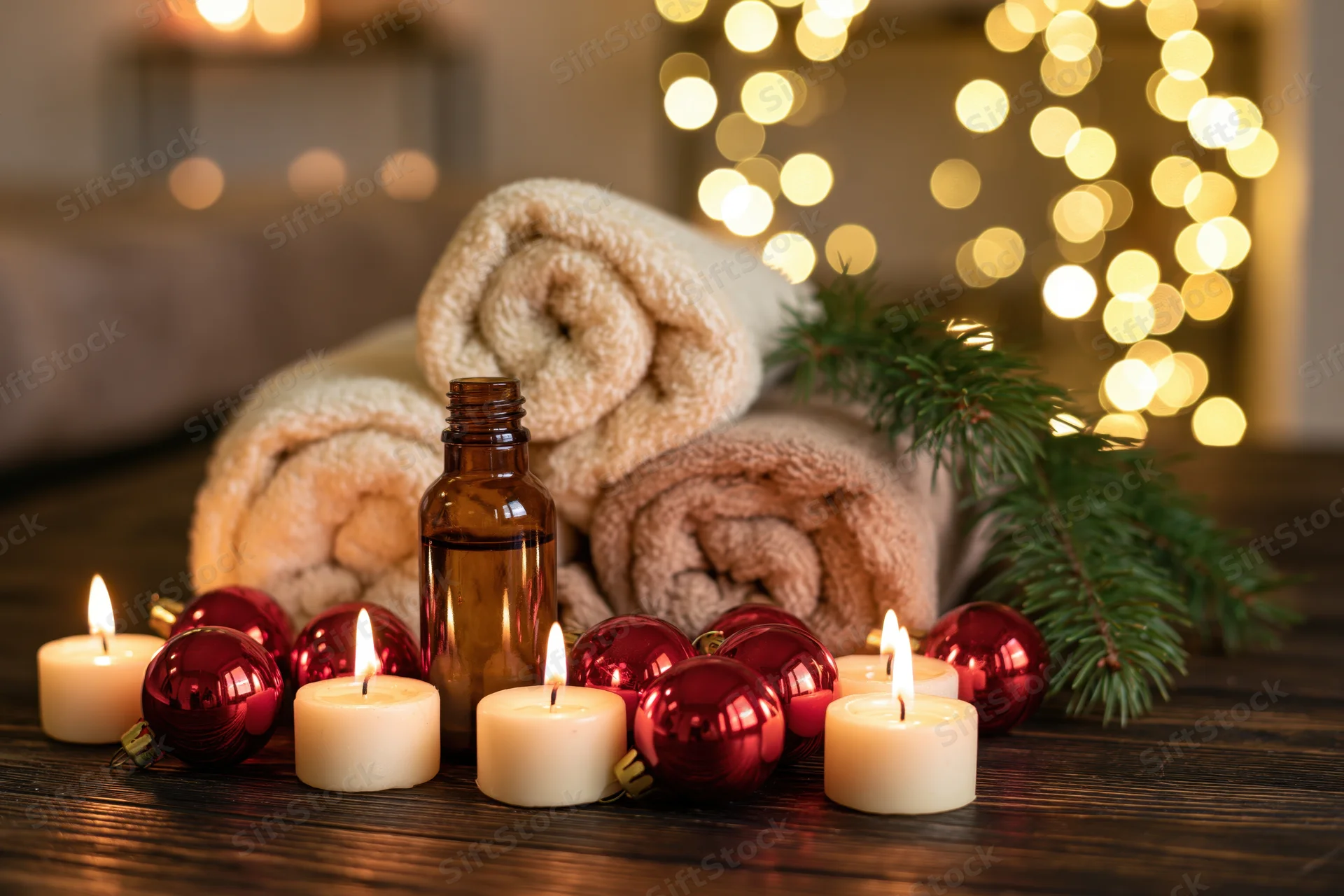 Spa still life with lit candles, rolled towels, essential oil bottle, red ornaments and pine sprig on dark wooden table with festive lights in background